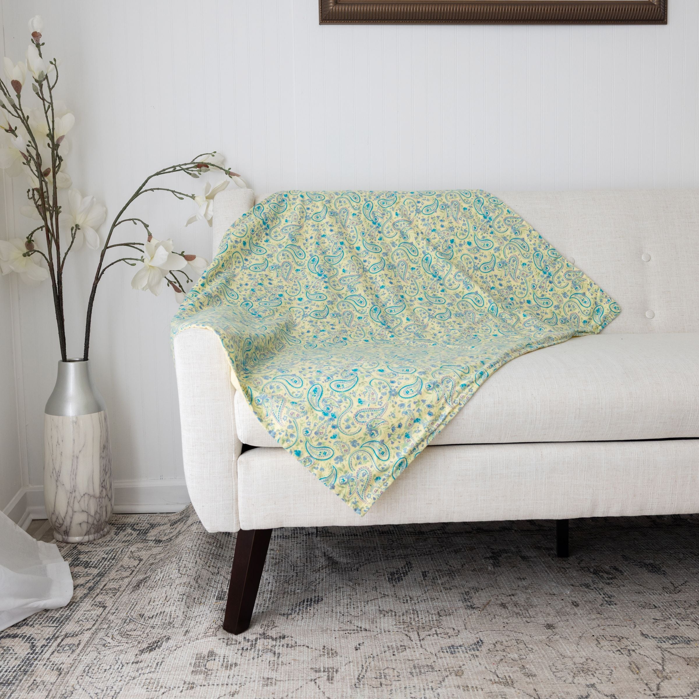Blue Toby cotton flannel blanket in a yellow and teal paisley pattern draped over a white sofa with a vase of flowers on a side table.