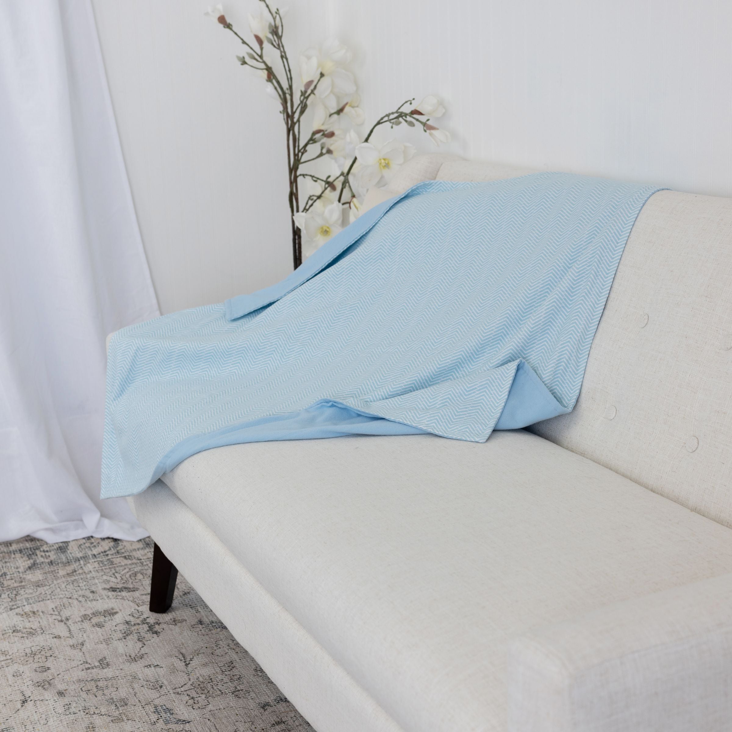 Blue Toby cotton flannel blanket in sky blue chevron pattern draped over a white sofa with a white curtain and plant in the background